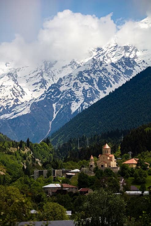 A mountain village of red-roofed houses and a church spire below pine forest and snow-covered peaks
